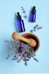 Bottles of essential oil, mortar and pestle with lavender on light blue background, flat lay