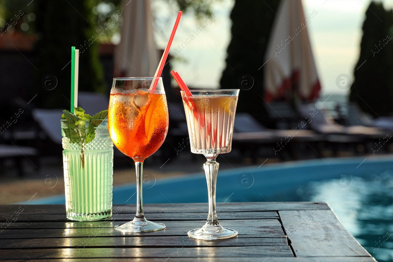 Photo of Glasses of fresh summer cocktails on wooden table near swimming pool outdoors