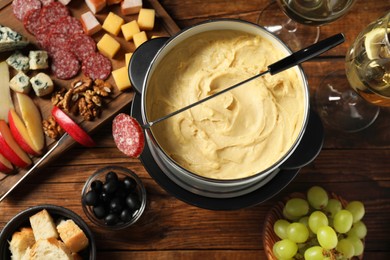 Photo of Fork with piece of sausage, melted cheese in fondue pot and other products on wooden table, flat lay