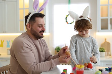 Photo of Easter celebration. Father with his little son painting eggs at white table in kitchen