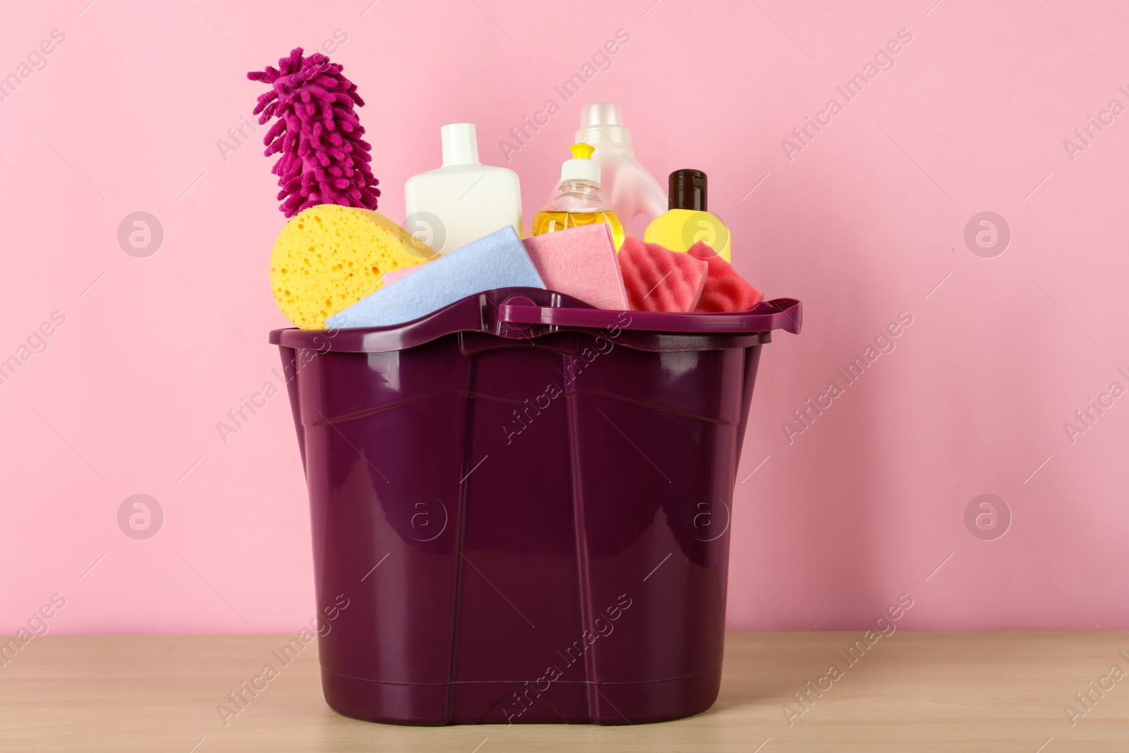 Photo of Bucket with different cleaning supplies on wooden floor near pink wall