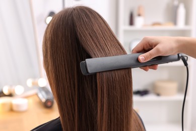 Photo of Hairdresser straightening woman's hair with flat iron in salon, closeup