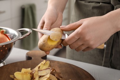 Photo of Woman peeling fresh potato with knife at light table indoors, closeup