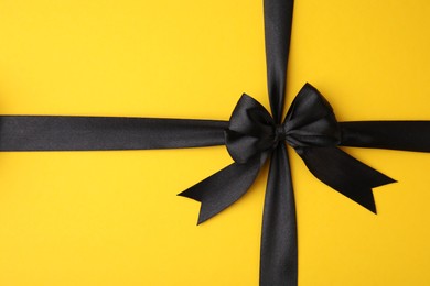 Photo of Black satin ribbon with bow on yellow background, top view