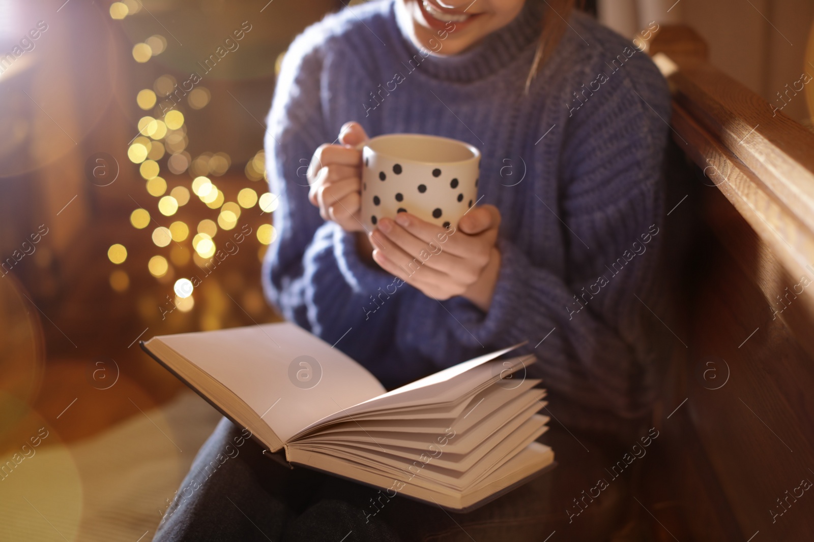 Photo of Woman with cup of hot beverage reading book at home in winter evening, closeup