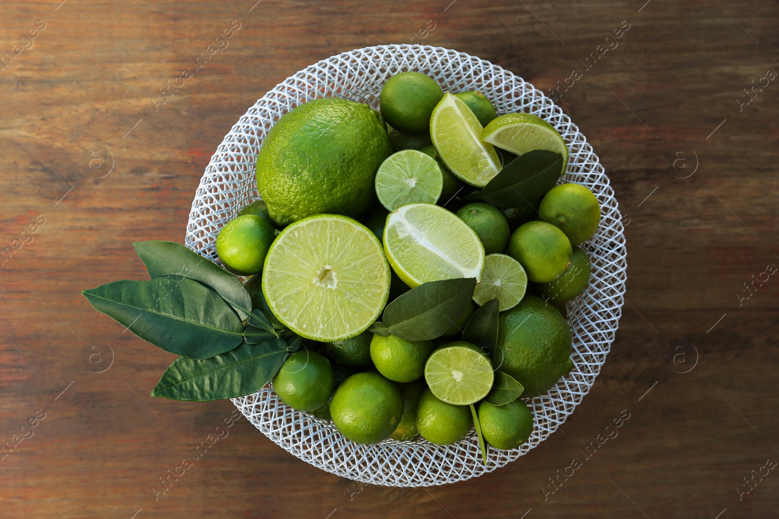 Photo of Fresh ripe limes and green leaves in bowl on wooden table, top view
