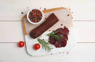 Delicious dry-cured beef basturma with rosemary and tomatoes on white wooden table, flat lay