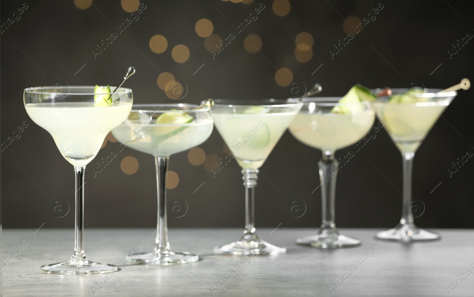 Photo of Glasses of martini with cucumber on grey table against blurred lights