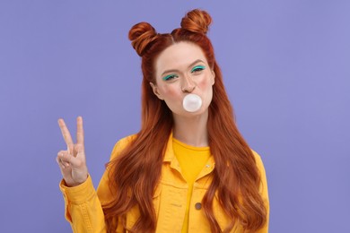 Photo of Portrait of beautiful woman with bright makeup blowing bubble gum and showing peace gesture on violet background