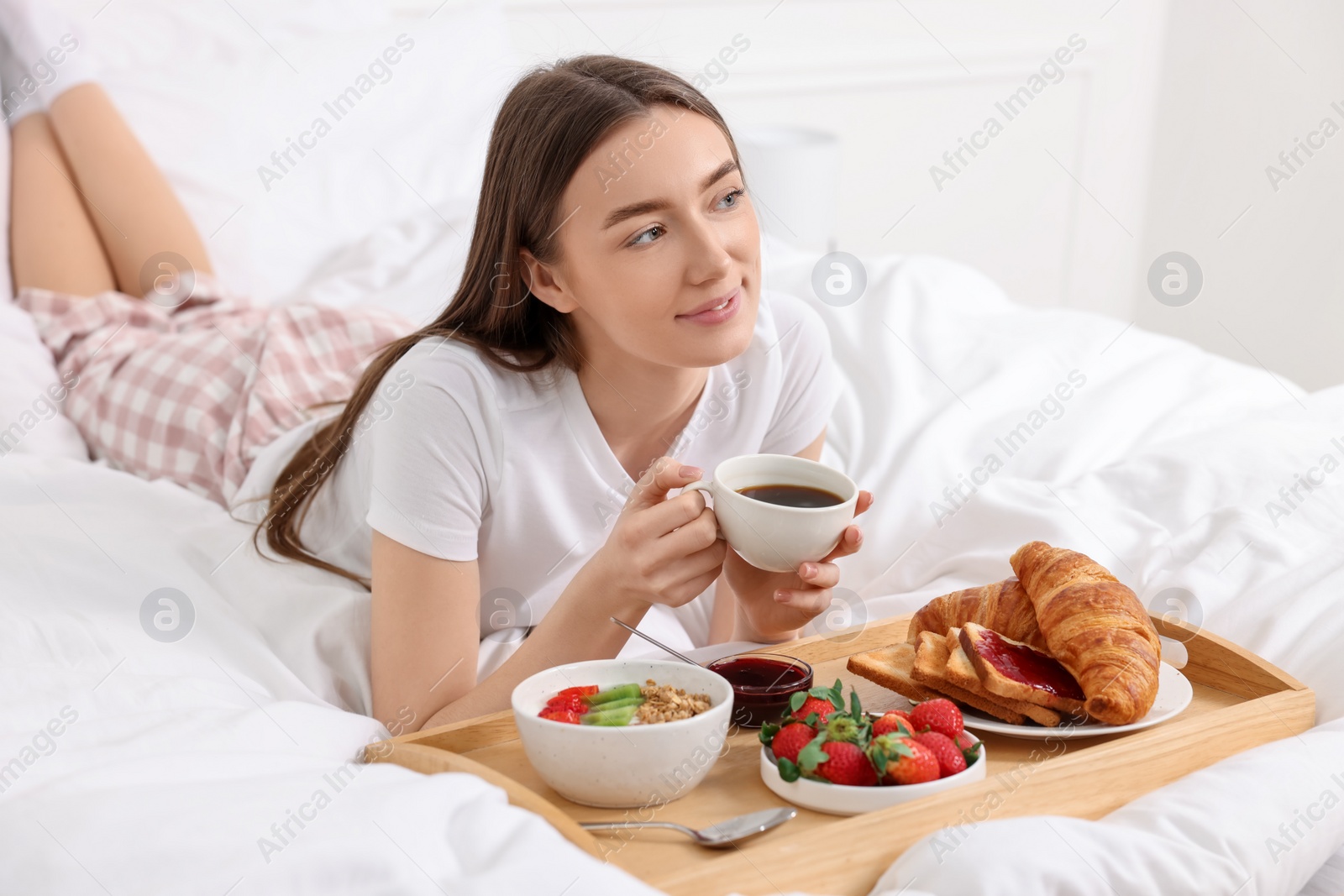 Photo of Beautiful woman drinking coffee near tray with breakfast on bed