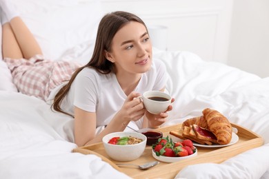 Beautiful woman drinking coffee near tray with breakfast on bed