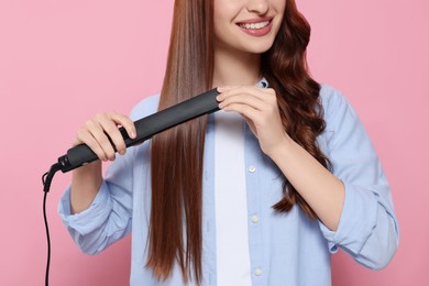 Young woman using hair iron on pink background, closeup
