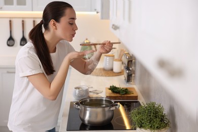 Beautiful woman with wooden spoon tasting soup in kitchen