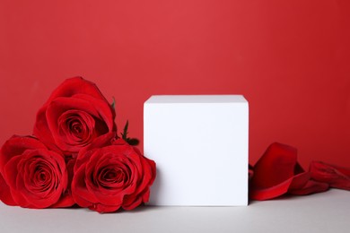 Photo of Stylish presentation for product. Cube, beautiful roses and petals on white table against red background