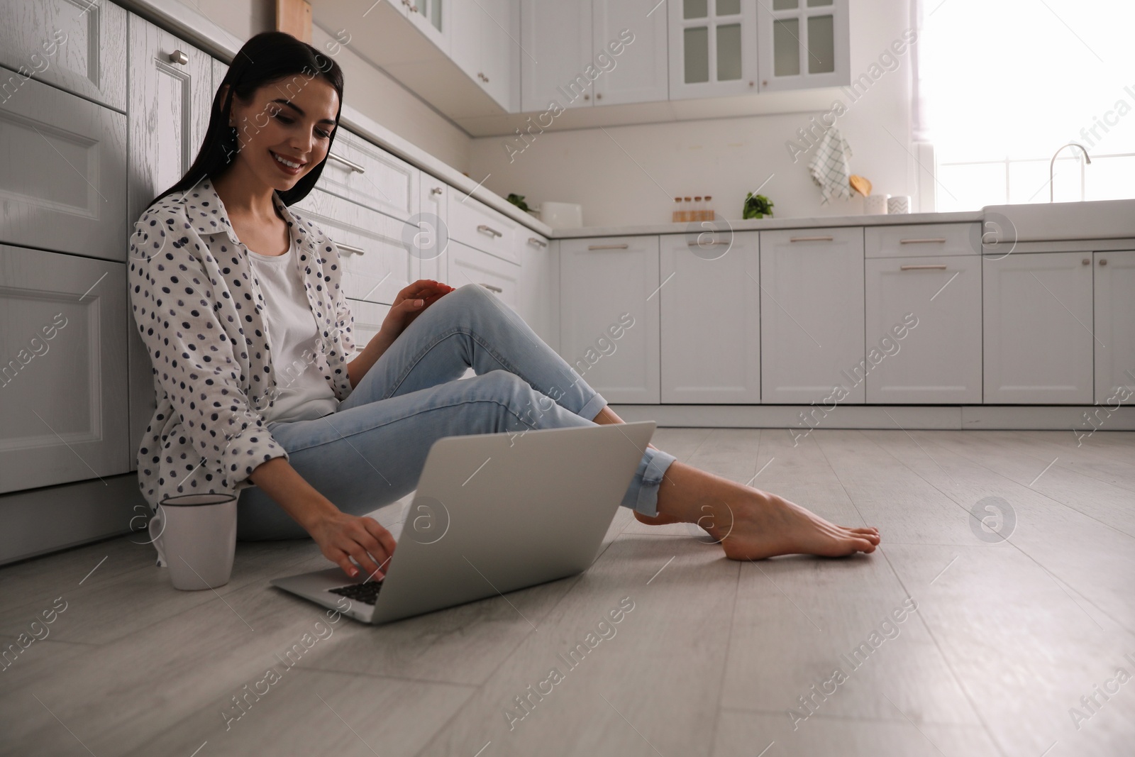 Photo of Happy woman with laptop and cup of drink sitting on warm floor in kitchen. Heating system