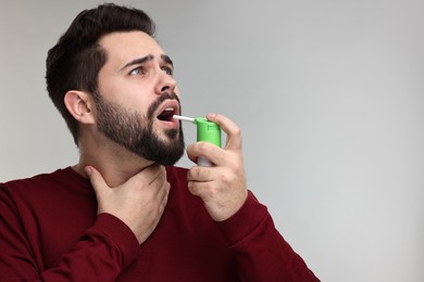 Young man using throat spray on grey background. Space for text