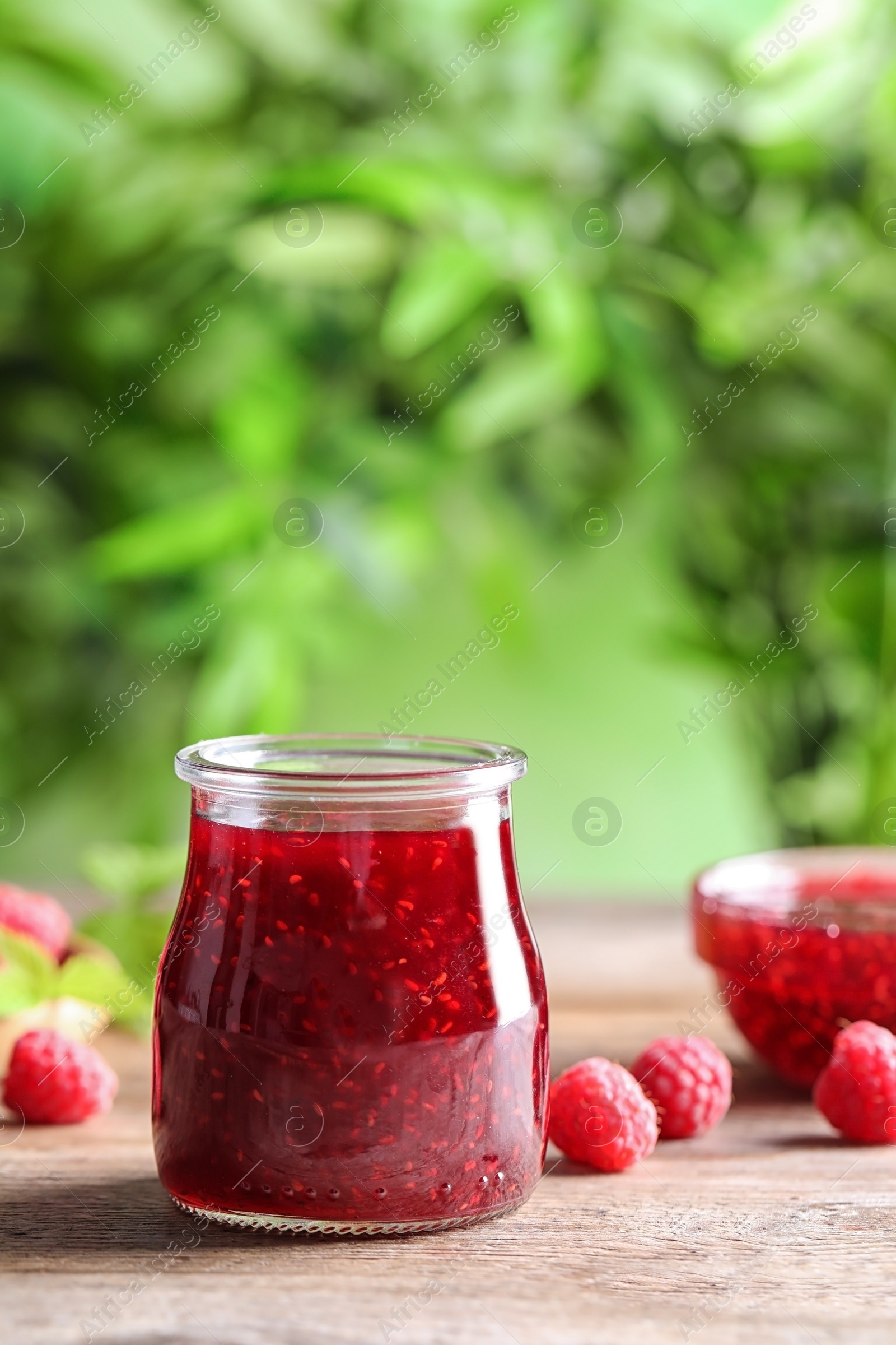 Photo of Glass jar of sweet jam with ripe raspberries on wooden table against blurred background