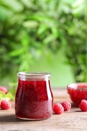 Photo of Glass jar of sweet jam with ripe raspberries on wooden table against blurred background