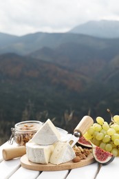 Delicious cheese, nuts and fruits on white wooden table against mountain landscape