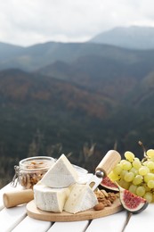 Photo of Delicious cheese, nuts and fruits on white wooden table against mountain landscape