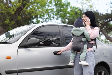 Photo of Mother holding her child in sling (baby carrier) while talking on smartphone near car outdoors