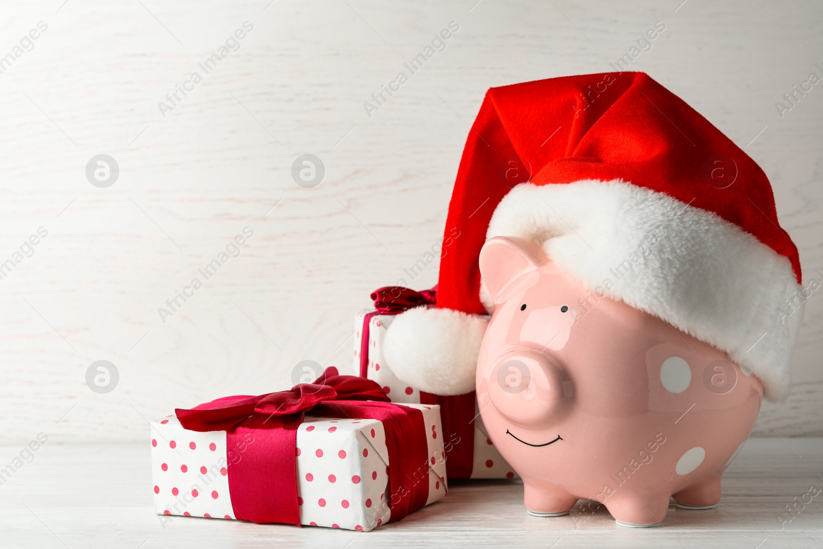 Photo of Piggy bank with Santa hat and gift boxes on white wooden table. Space for text