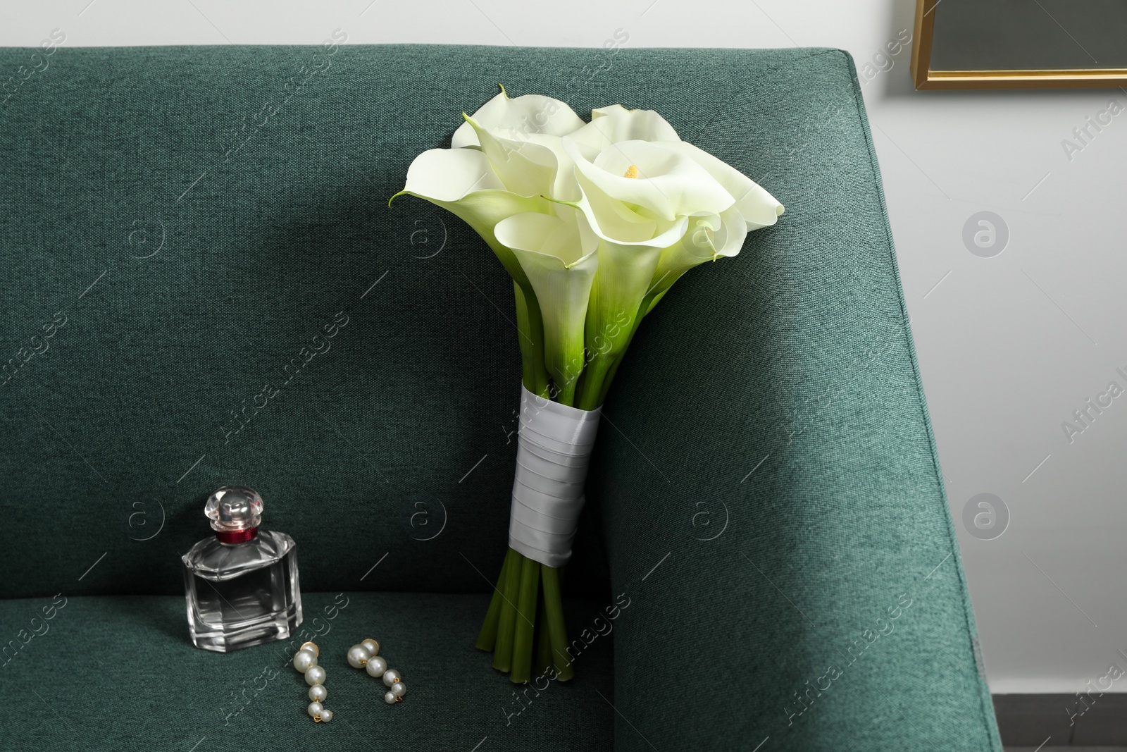Photo of Beautiful calla lily flowers tied with ribbon, bottle of perfume and earrings on sofa indoors