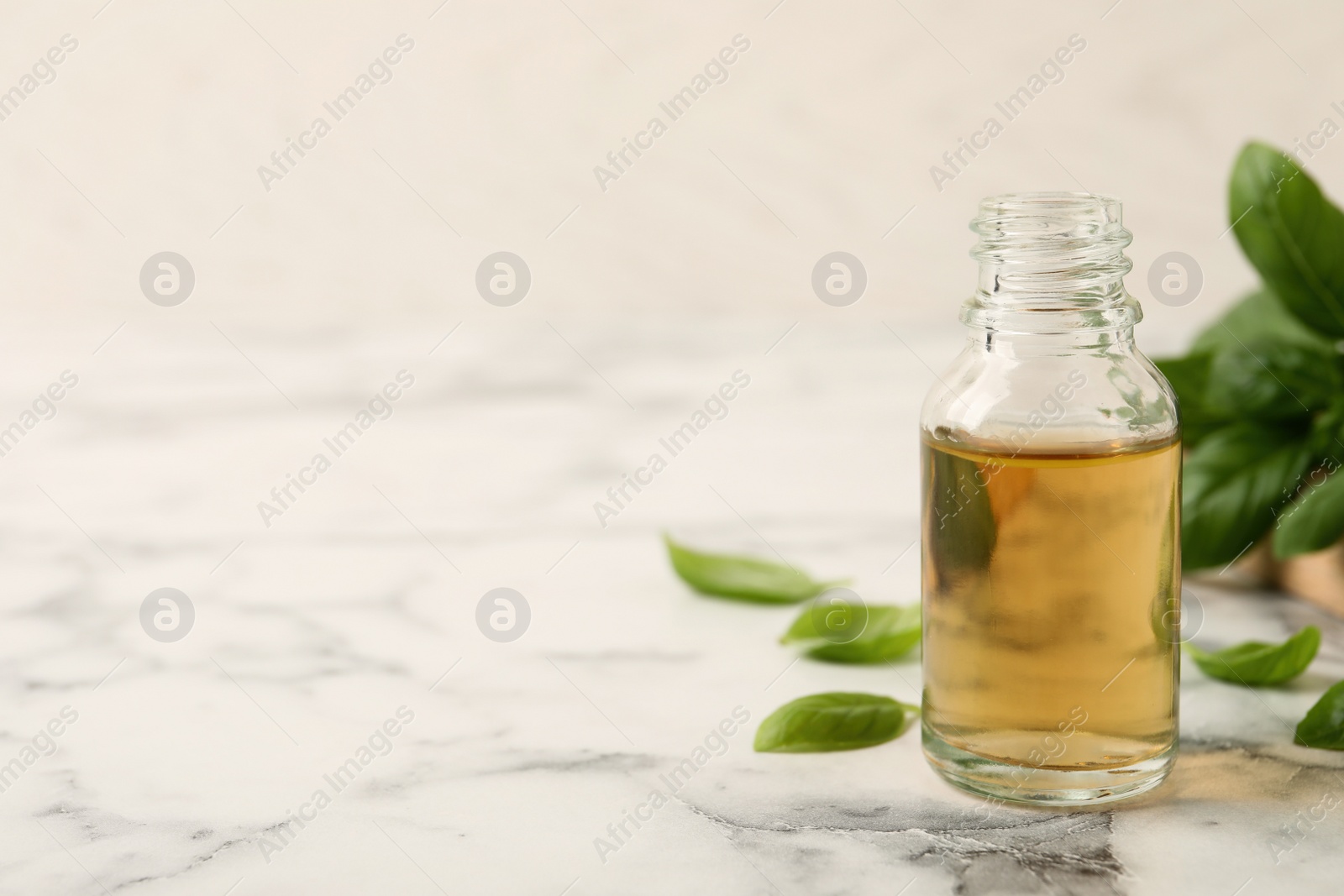 Photo of Glass bottle of basil essential oil and leaves on white marble table. Space for text