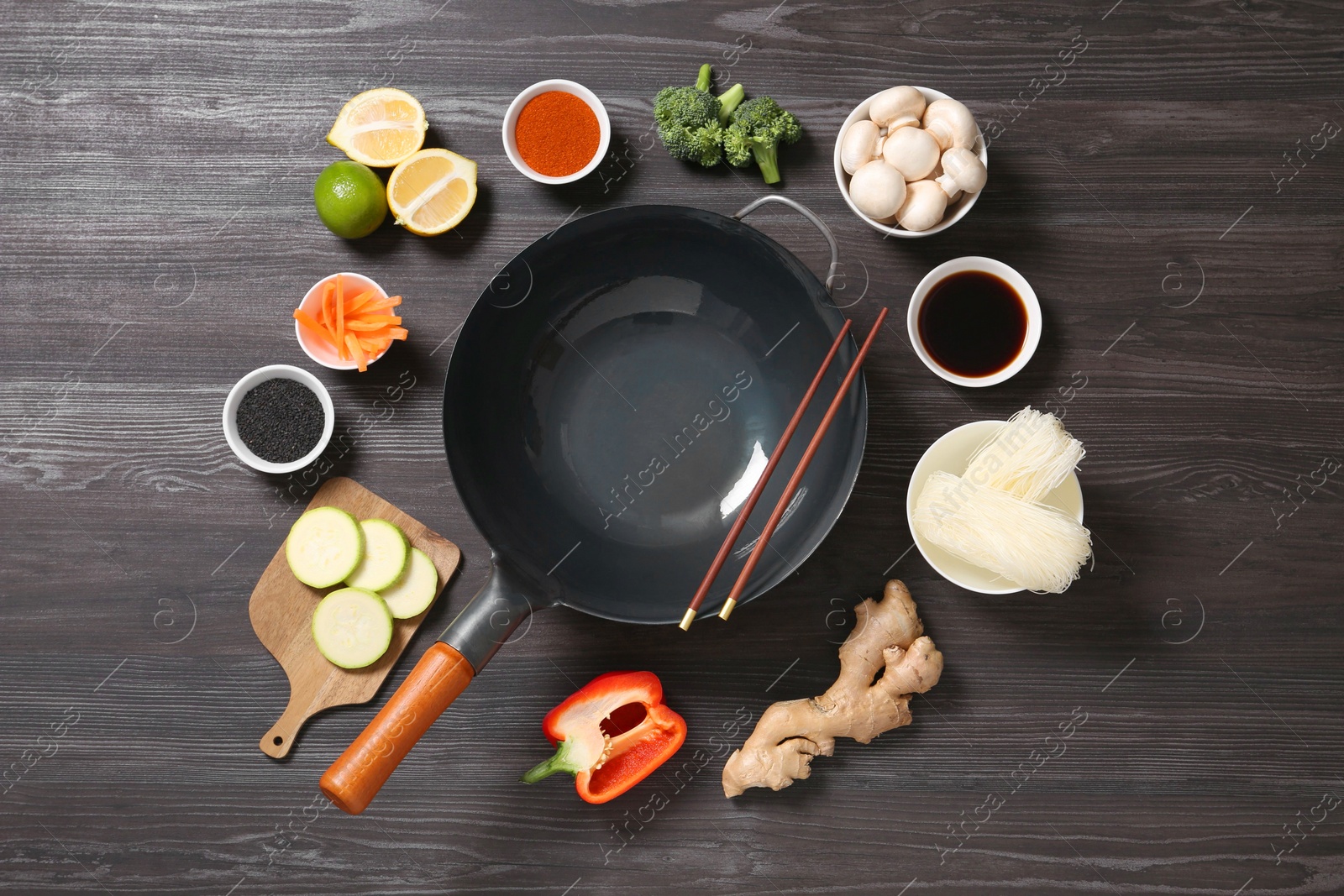 Photo of Empty iron wok and chopsticks surrounded by ingredients on dark grey wooden table, flat lay