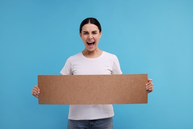 Photo of Angry woman holding blank cardboard banner on light blue background, space for text