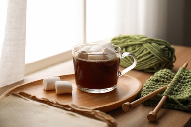 Photo of Cup of hot winter drink with marshmallows and knitting on wooden sill near window