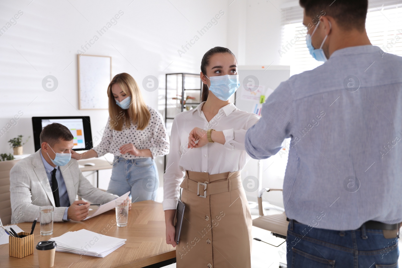 Photo of Coworkers with protective masks making elbow bump in office. Informal greeting during COVID-19 pandemic