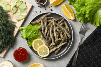 Delicious fried anchovies with lemon, lettuce leaves and sauces served on light grey table, flat lay