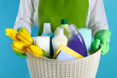 Spring cleaning. Woman holding basket with detergents, flowers and rags on light blue background, closeup