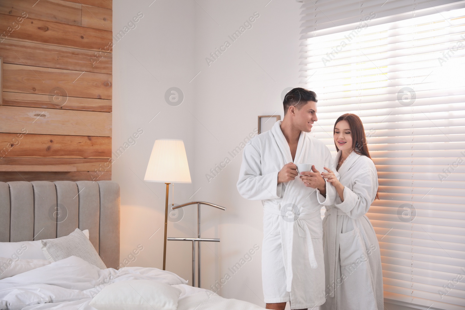 Photo of Happy couple wearing bathrobes near window in bedroom