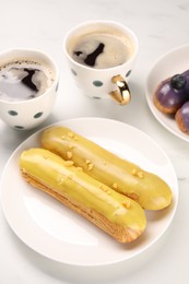 Tasty glazed eclairs and cups of coffee on white table, closeup