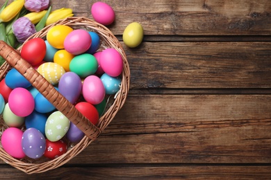 Photo of Colorful Easter eggs in wicker basket and tulips on wooden table, flat lay. Space for text