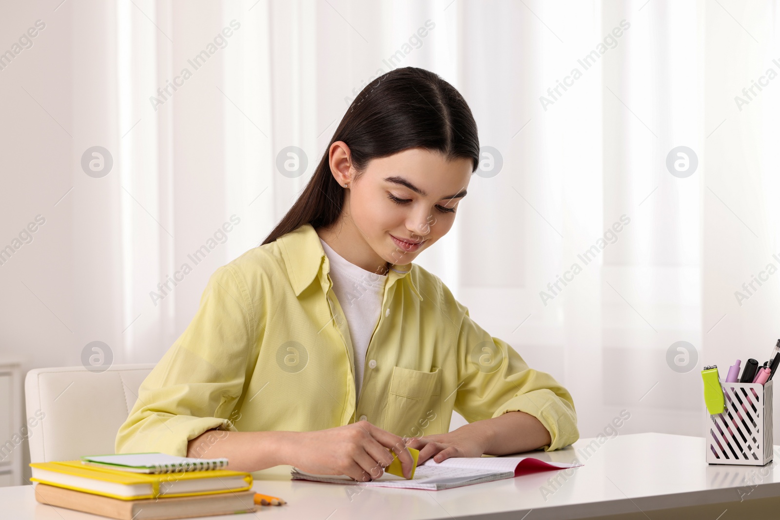 Photo of Teenage girl erasing mistake in her notebook at white desk indoors