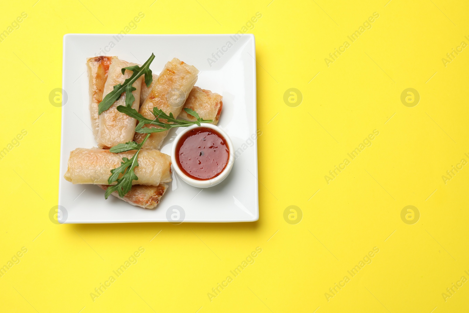 Photo of Fried spring rolls, arugula and sauce on yellow table, top view. Space for text