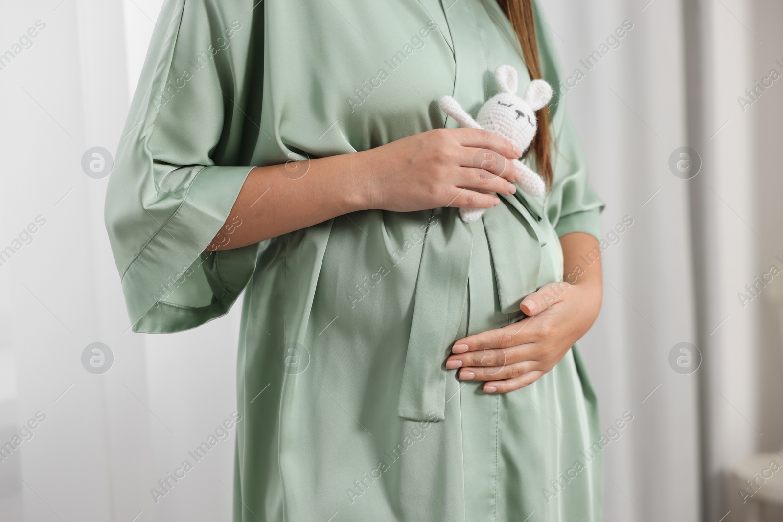 Photo of Pregnant woman in green dressing gown with bunny toy near window indoors, closeup