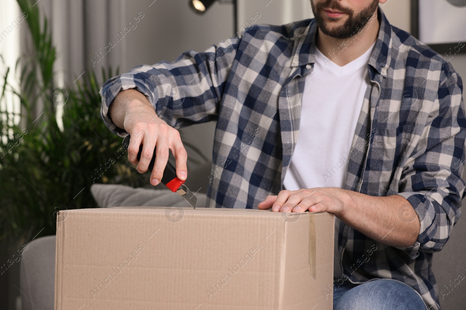 Photo of Man using utility knife to open parcel at home, closeup