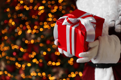 Santa Claus holding Christmas gift against blurred festive lights, closeup