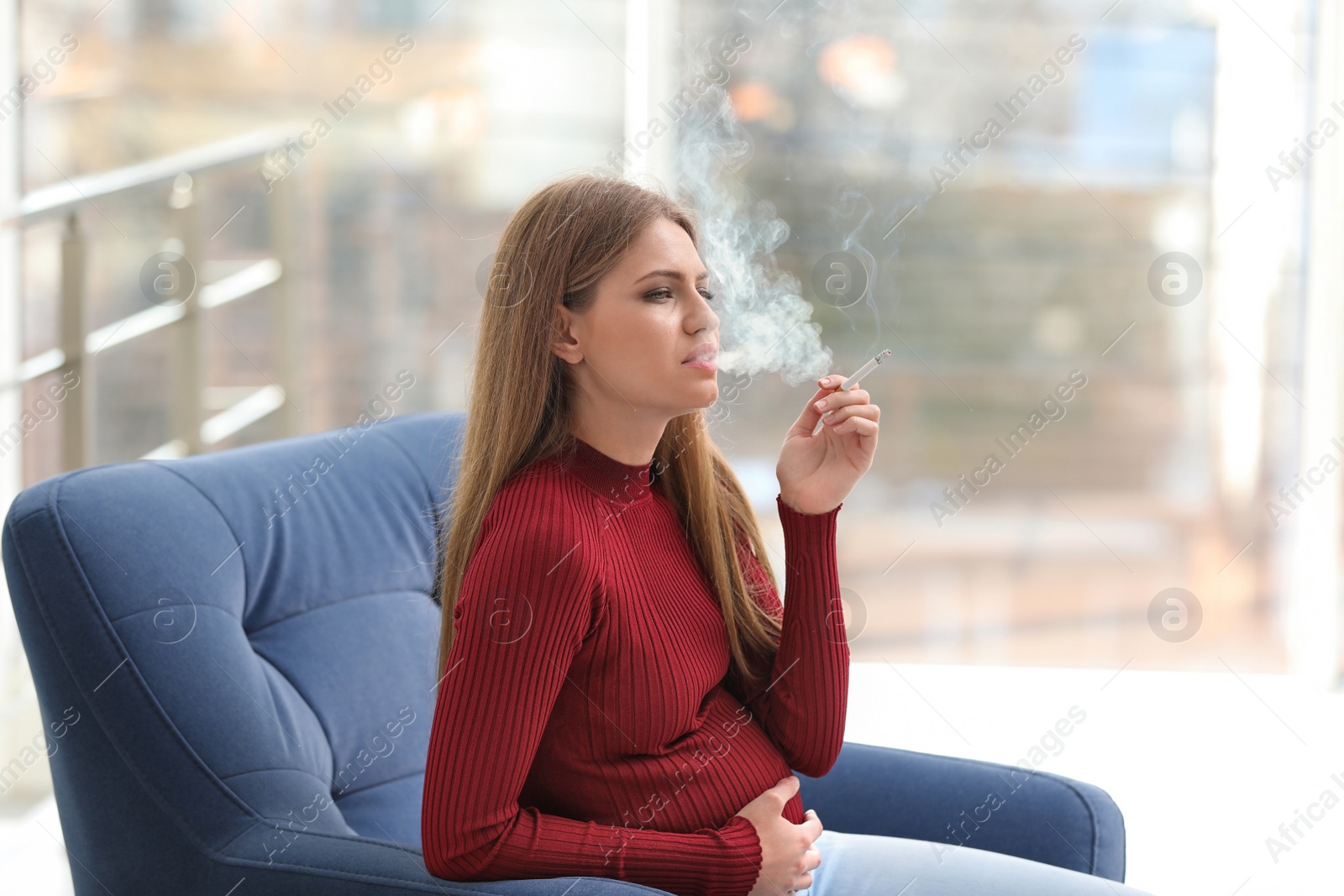 Photo of Young pregnant woman smoking cigarette at home. Harm to unborn baby