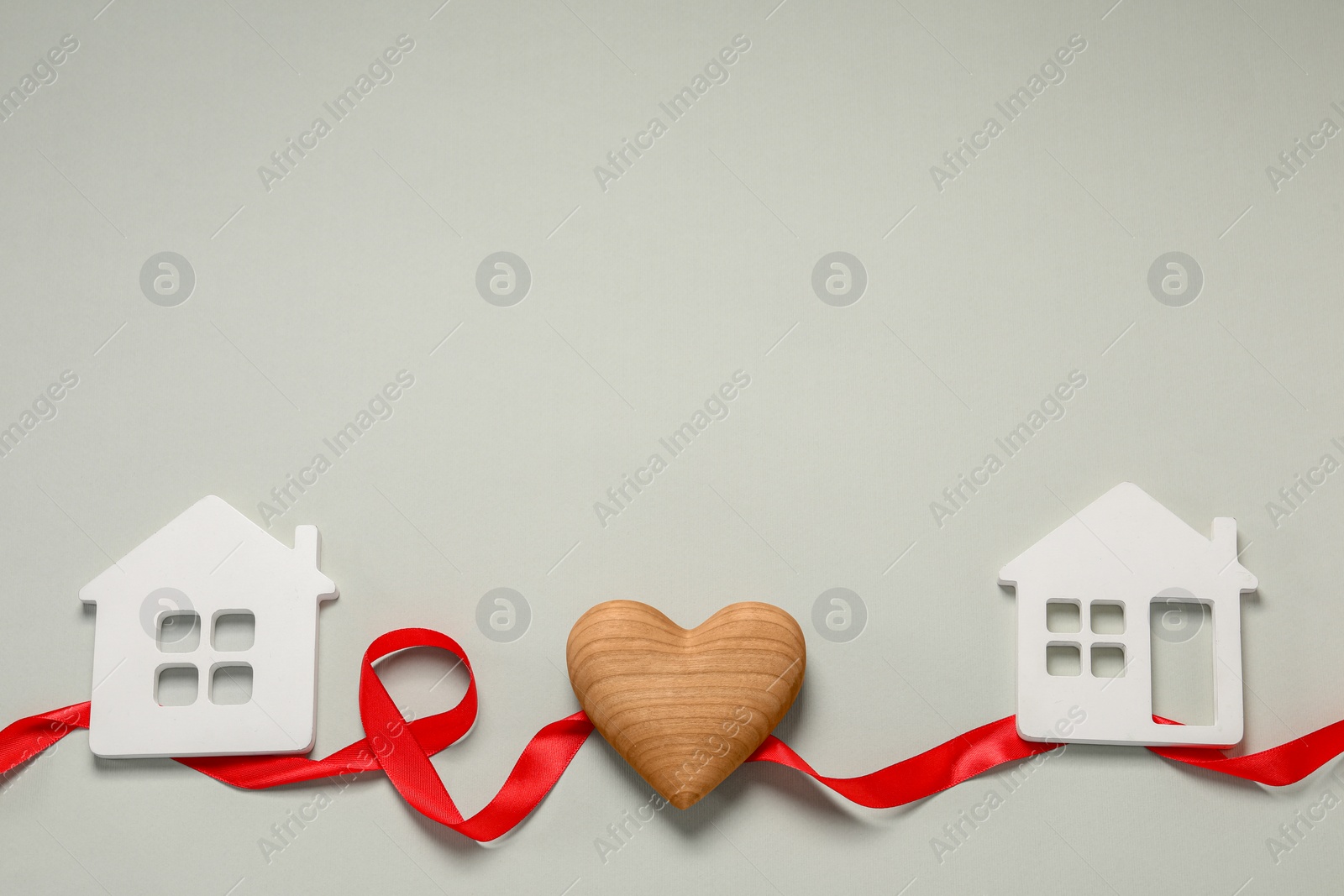 Photo of Red ribbon and decorative heart between two house models on light gray background symbolizing connection in long-distance relationship, above view. Space for text