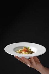 Photo of Woman holding plate of tasty spaghetti with prosciutto and microgreens on black background, closeup and space for text. Exquisite presentation of pasta dish