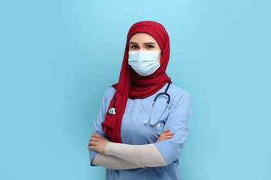 Photo of Muslim woman wearing hijab, medical uniform and protective mask on light blue background