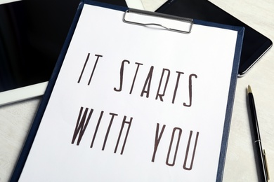 Photo of Clipboard with phrase It Starts With You, tablet and smartphone on white table, closeup