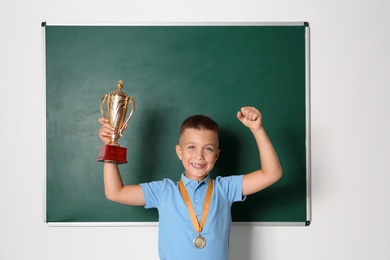 Photo of Happy boy with golden winning cup and medal near chalkboard in classroom