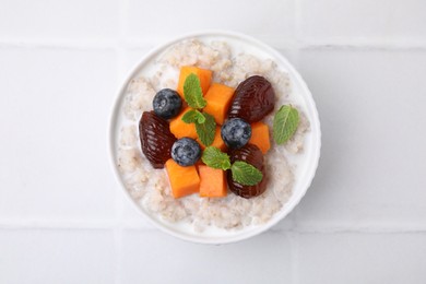 Delicious barley porridge with blueberries, pumpkin, dates and mint in bowl on white tiled table, top view