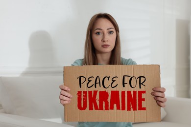 Image of Unhappy young woman holding sign with phrase Peace for Ukraine on sofa indoors